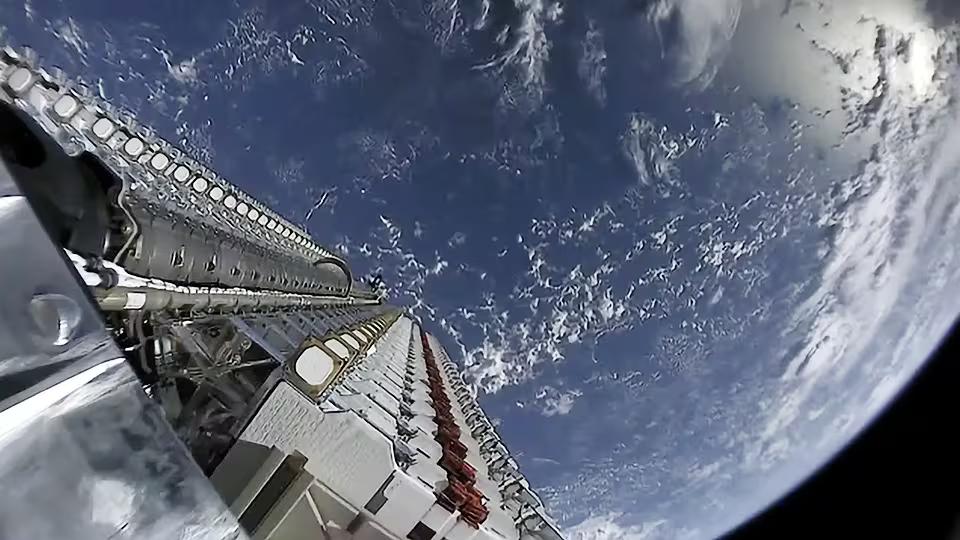 Fisheye view from a Falcon 9 second stage looking back at a stack of 60 Starlink satellites still folded together, with the cloud-streaked Earth filling the right of the frame just before deployment.