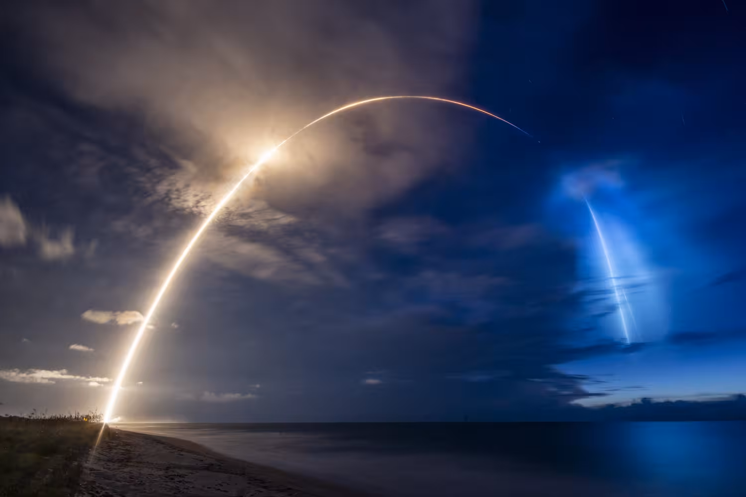 Long-exposure photograph of a Falcon 9 rocket launching at twilight from Cape Canaveral, the orange ascent trail arching over the Atlantic and the blue plume of the returning first stage glowing in the upper atmosphere alongside it